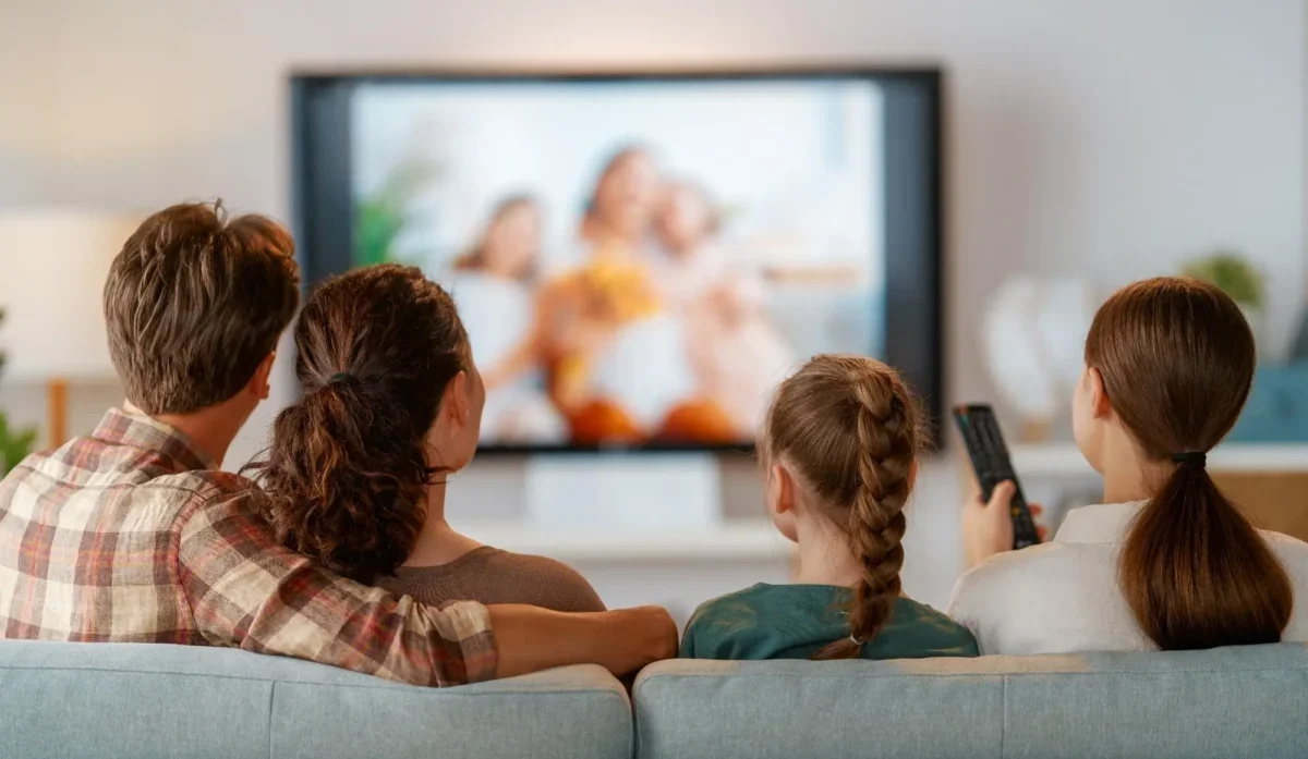 Family seated on a sofa watching a blurry TV show together, one person holding a remote control to the right