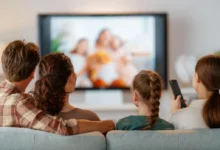 Family seated on a sofa watching a blurry TV show together, one person holding a remote control to the right