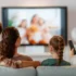Family seated on a sofa watching a blurry TV show together, one person holding a remote control to the right