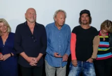 Group of six adults posing for a photo at an event, wearing casual outfits and standing shoulder to shoulder against a plain backdrop.
