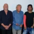 Group of six adults posing for a photo at an event, wearing casual outfits and standing shoulder to shoulder against a plain backdrop.