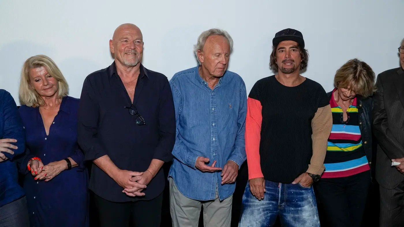 Group of six adults posing for a photo at an event, wearing casual outfits and standing shoulder to shoulder against a plain backdrop.