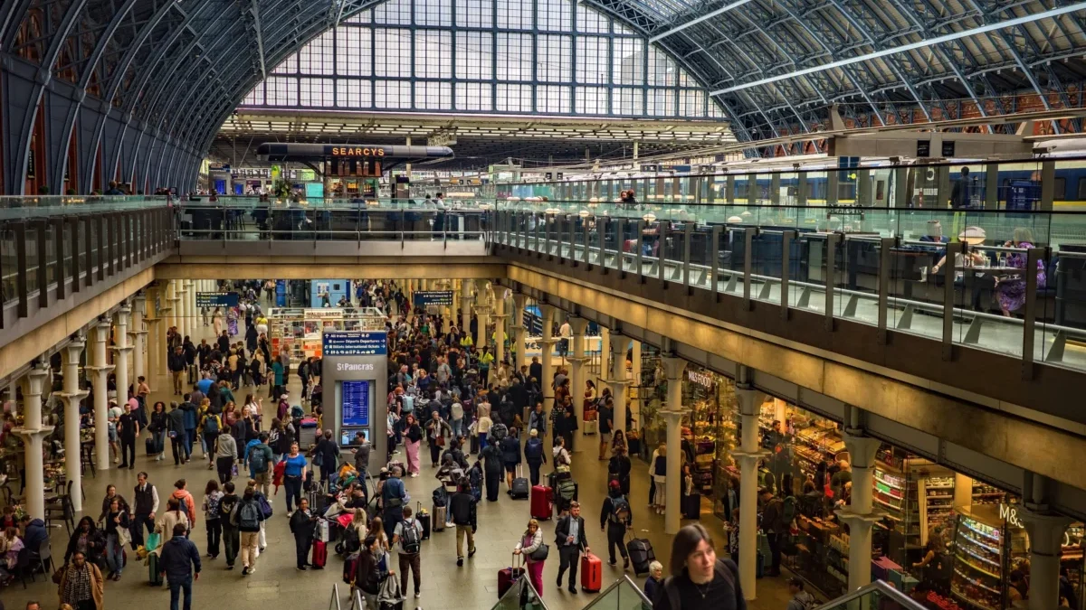 Busy train station concourse filled with travelers and luggage beneath a glass vaulted roof.