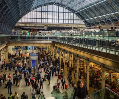 Busy train station concourse filled with travelers and luggage beneath a glass vaulted roof.