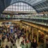 Busy train station concourse filled with travelers and luggage beneath a glass vaulted roof.