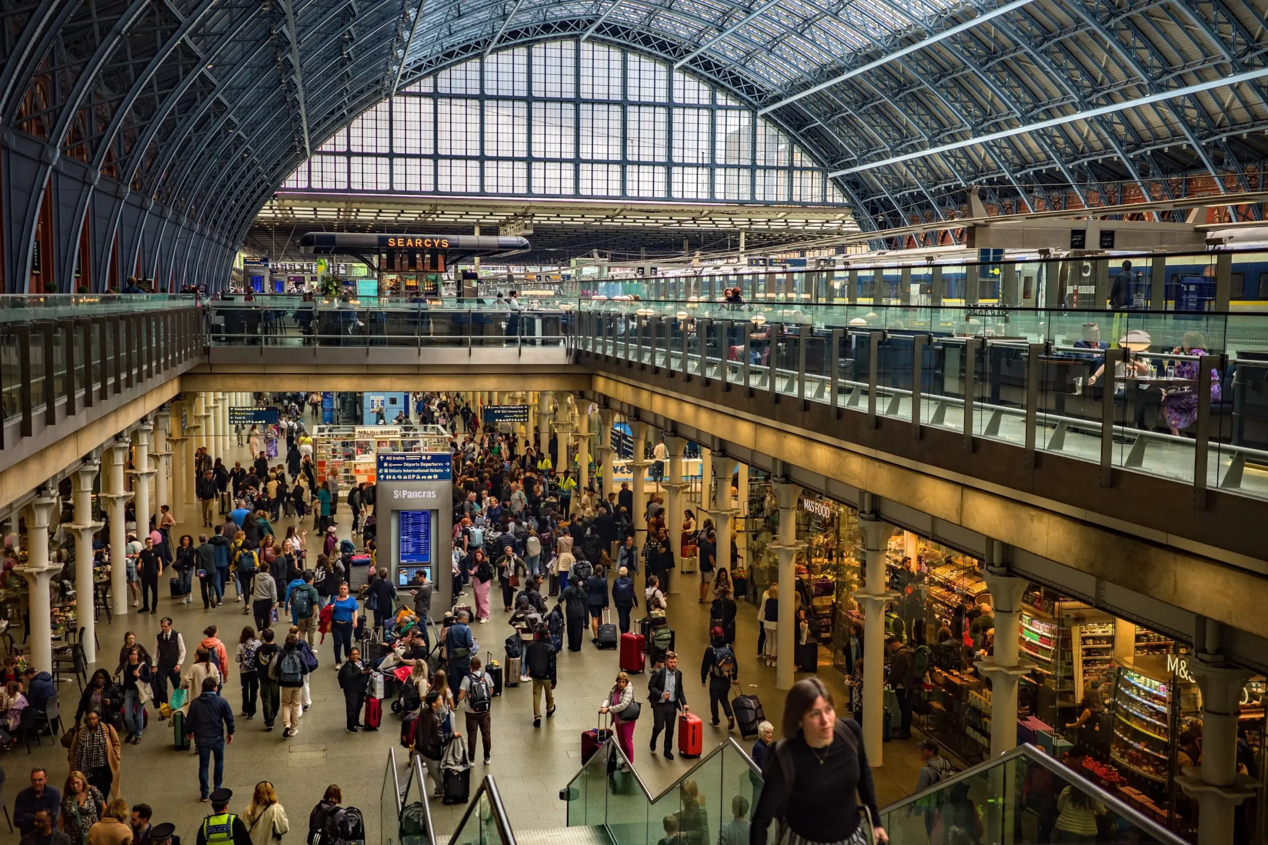 Busy train station concourse filled with travelers and luggage beneath a glass vaulted roof.