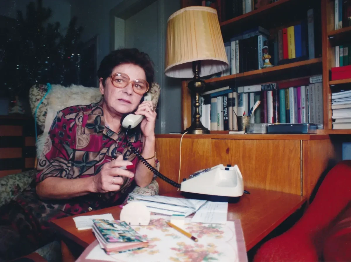 Jiřina Švorcová sedí u stolu a telefonuje.