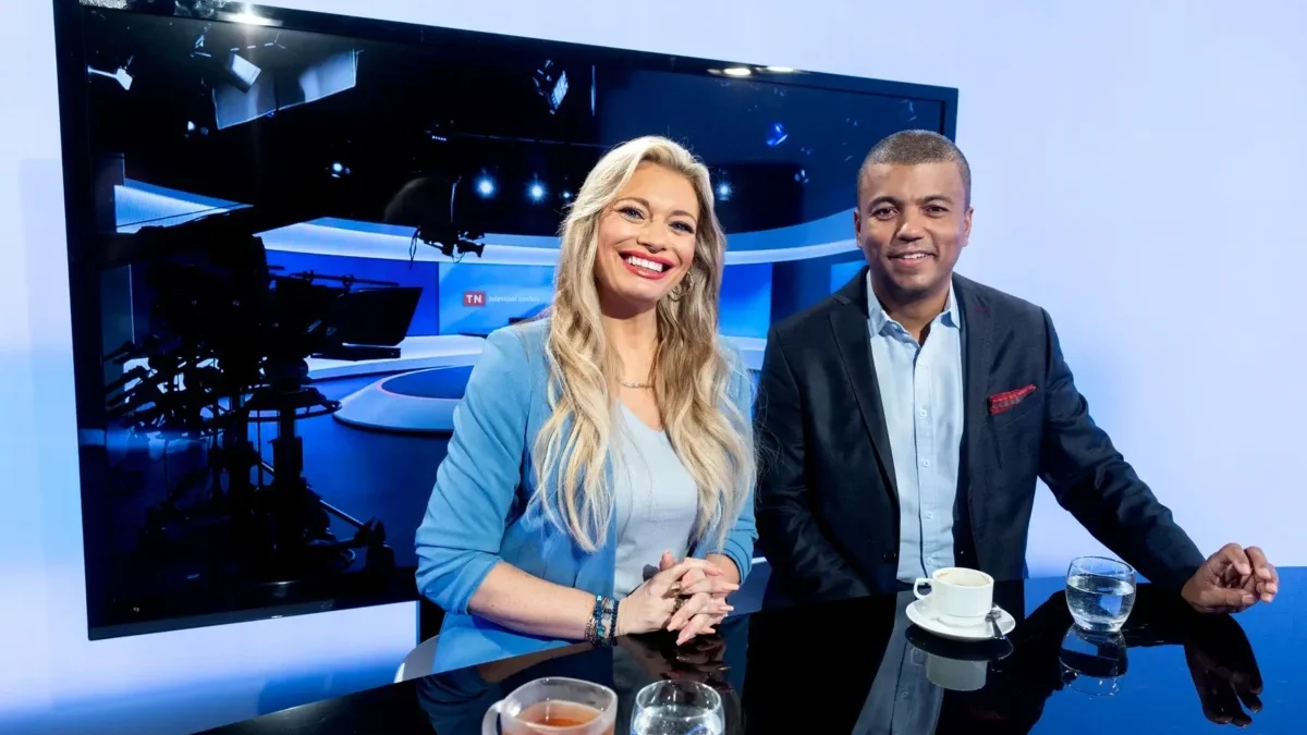 Two TV hosts smile at a glossy studio desk, with a large monitor and studio lights behind them and coffee cups on the table.
