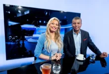 Two TV hosts smile at a glossy studio desk, with a large monitor and studio lights behind them and coffee cups on the table.