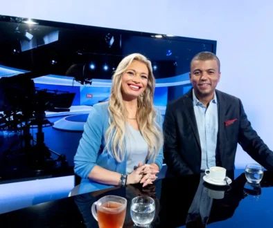 Two TV hosts smile at a glossy studio desk, with a large monitor and studio lights behind them and coffee cups on the table.