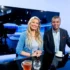 Two TV hosts smile at a glossy studio desk, with a large monitor and studio lights behind them and coffee cups on the table.