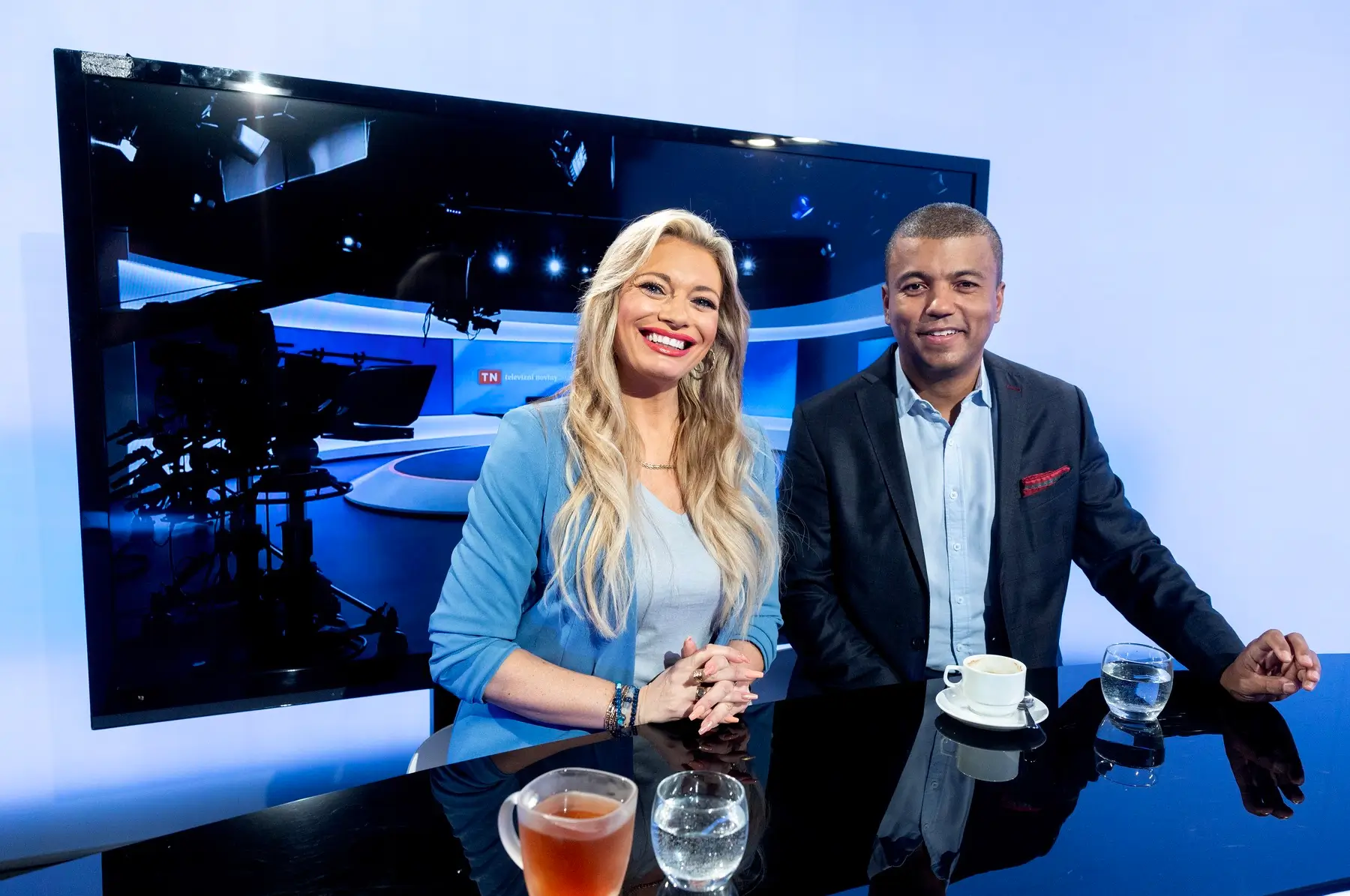 Two TV hosts smile at a glossy studio desk, with a large monitor and studio lights behind them and coffee cups on the table.