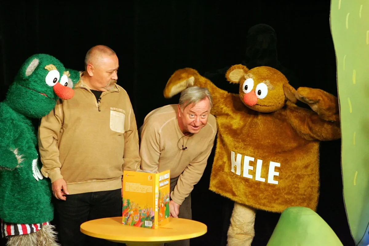 profimedia-0157566689 1 | TVGURU.cz Two men in beige tops stand with colorful fuzzy monsters on a stage, a green monster on the left and a brown monster on the right, engaging with a table and a book model.