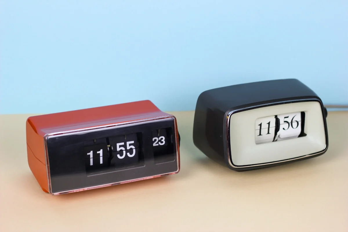 Two vintage flip clocks on a pale desk, showing times 11:55 (orange clock) and 11:56 (black clock) against a light blue wall.