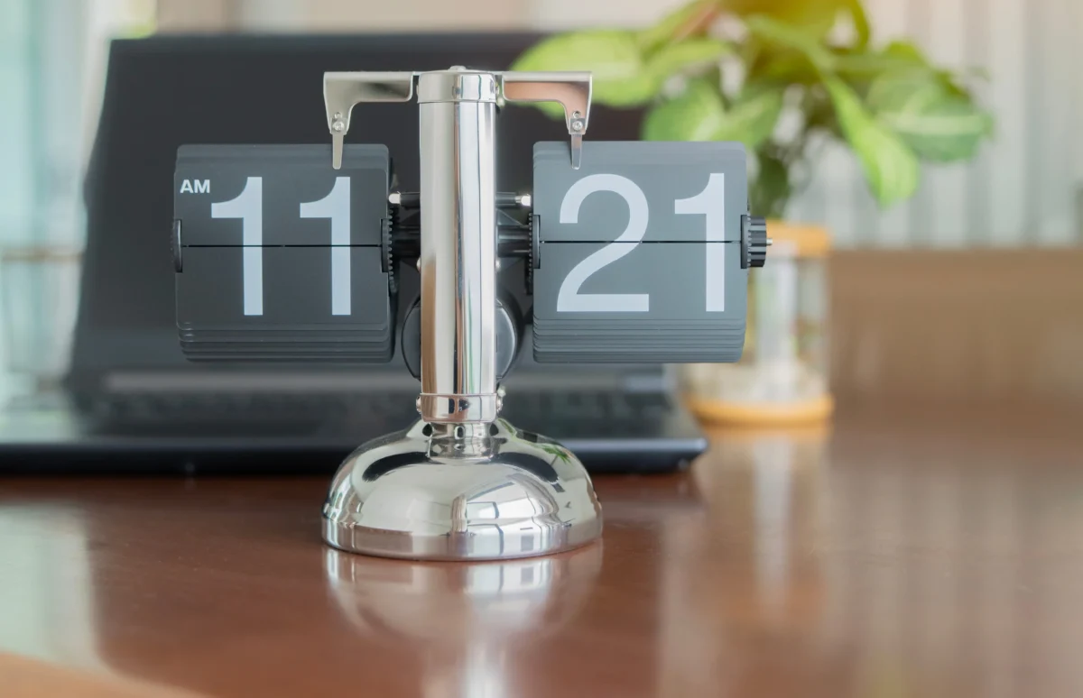 Retro flip clock on a desk showing 11:21 AM, with a laptop and green plant blurred in the background.