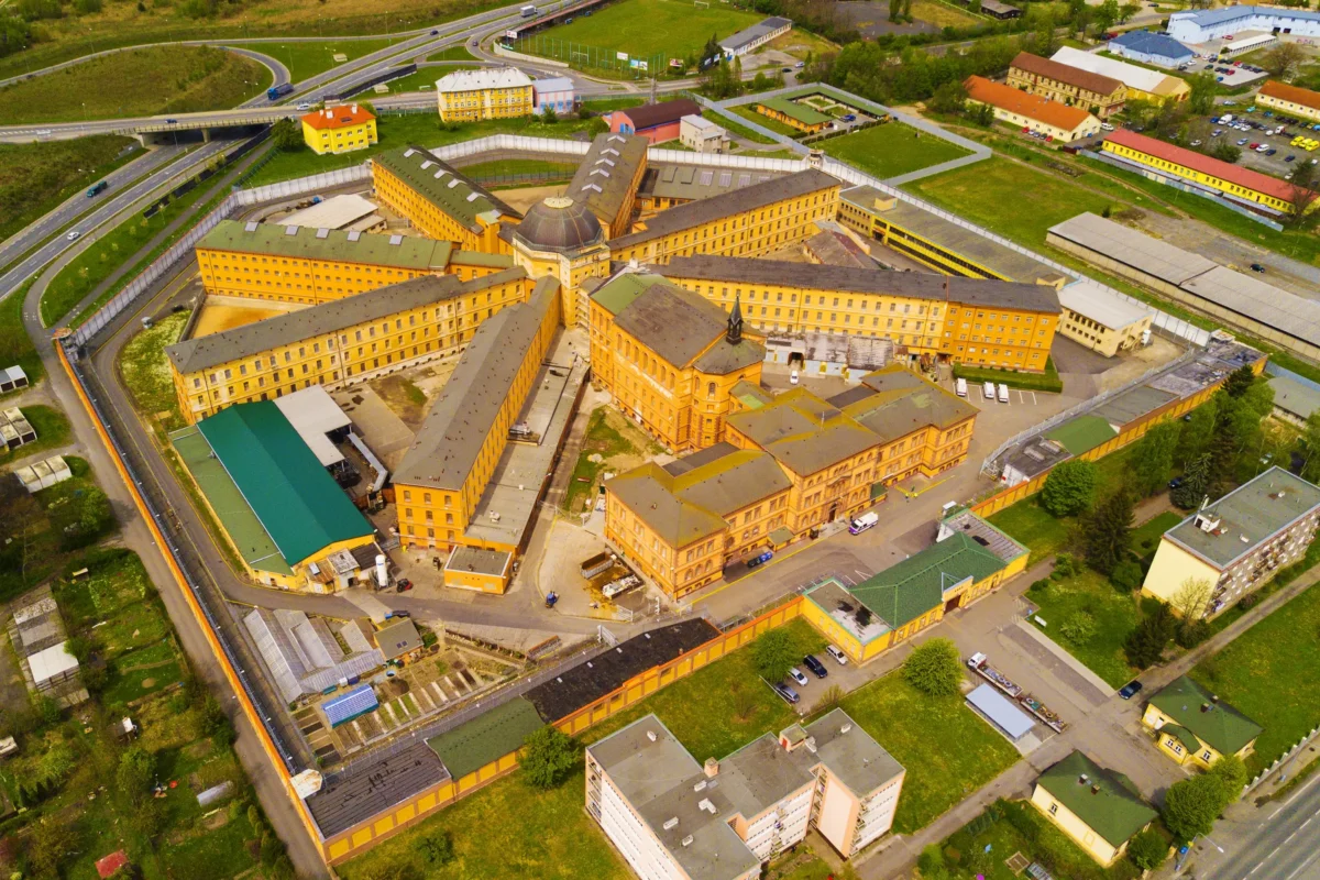 shutterstock_633642842 1 | TVGURU.cz Aerial view of a large yellow brick campus arranged in a star-shaped layout around a central domed building, with roads and green spaces nearby.