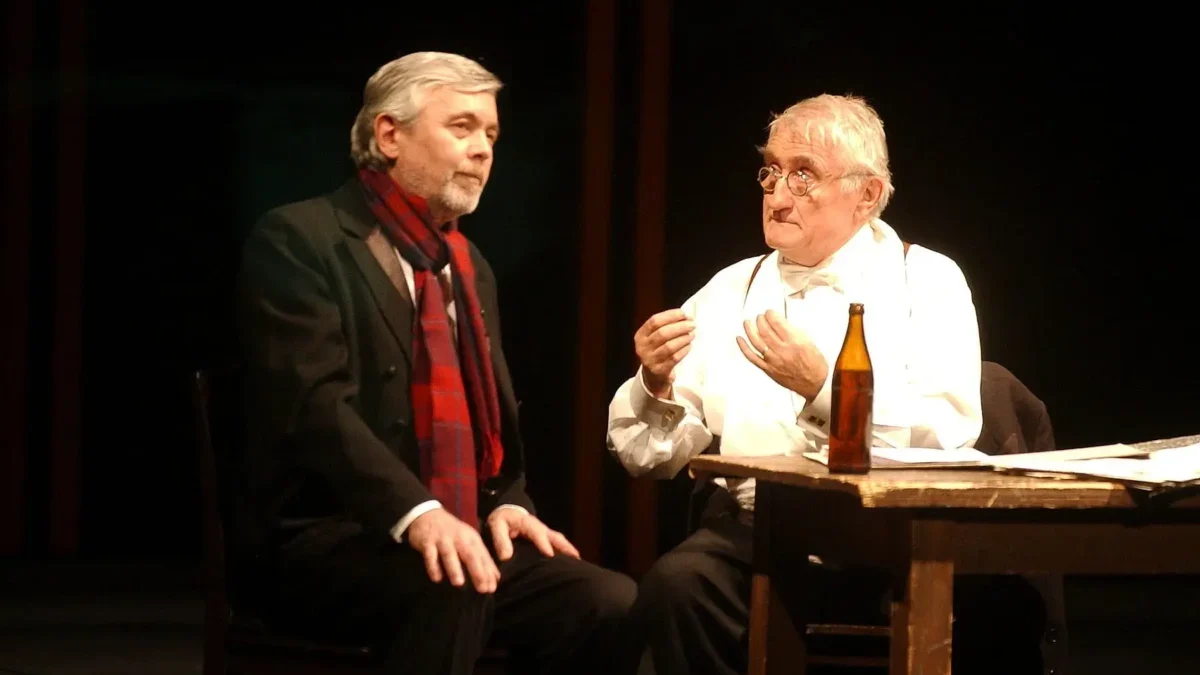 Stage dialogue: two men seated at a wooden table on dimly lit set, one in a dark suit with a red scarf and the other in white with suspenders and glasses, a bottle on the table