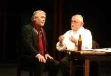 Stage dialogue: two men seated at a wooden table on dimly lit set, one in a dark suit with a red scarf and the other in white with suspenders and glasses, a bottle on the table