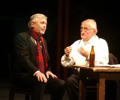 Stage dialogue: two men seated at a wooden table on dimly lit set, one in a dark suit with a red scarf and the other in white with suspenders and glasses, a bottle on the table