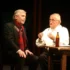 Stage dialogue: two men seated at a wooden table on dimly lit set, one in a dark suit with a red scarf and the other in white with suspenders and glasses, a bottle on the table