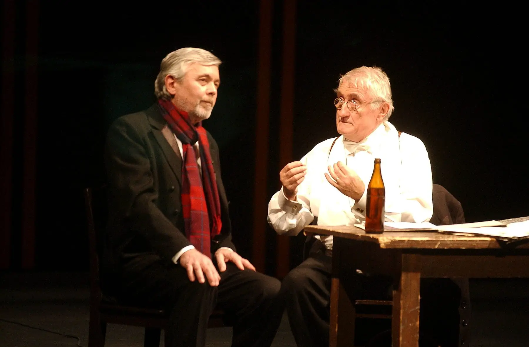 Stage dialogue: two men seated at a wooden table on dimly lit set, one in a dark suit with a red scarf and the other in white with suspenders and glasses, a bottle on the table