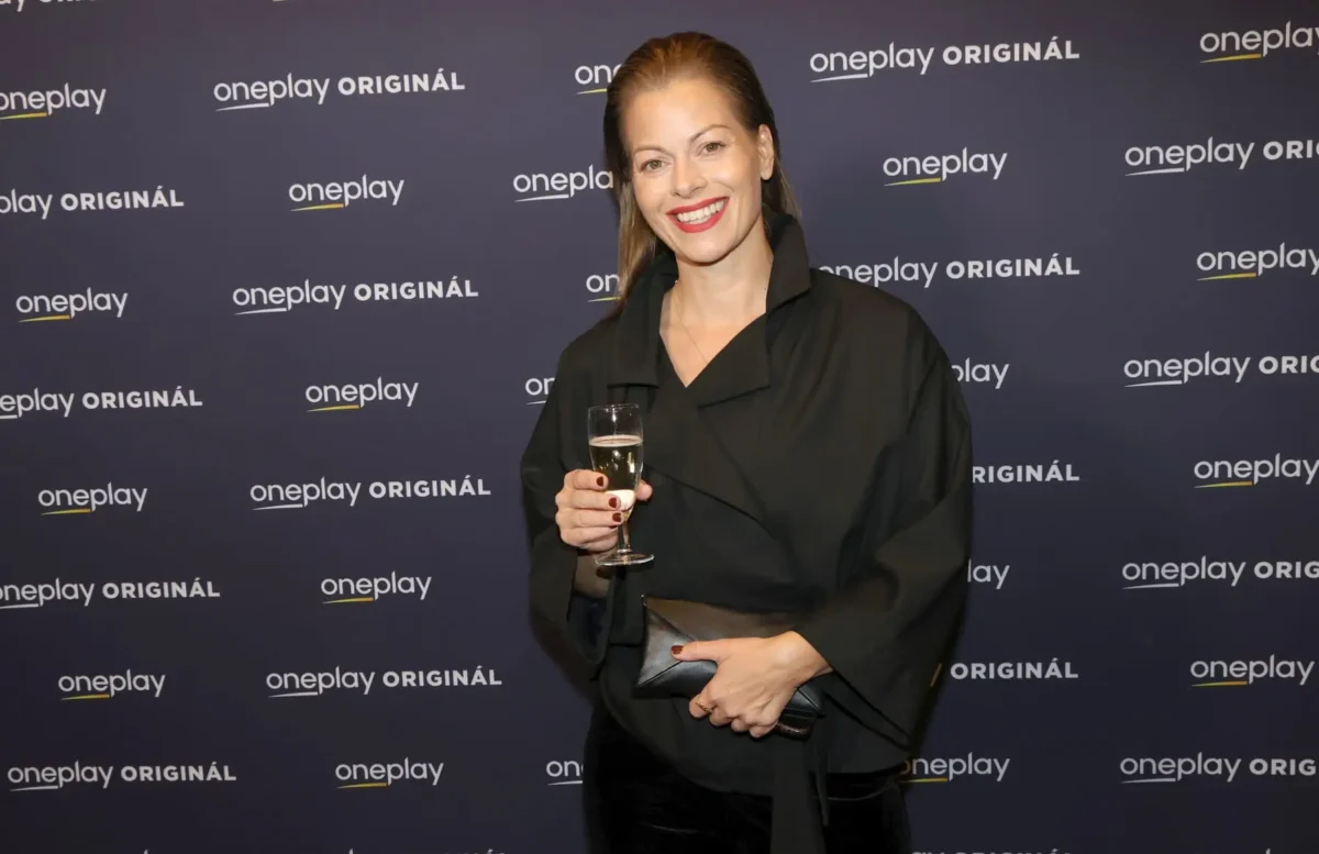 profimedia-1097014331 1 | TVGURU.cz Smiling woman in a black outfit holding a champagne flute, posing in front of a OnePlay Original backdrop.