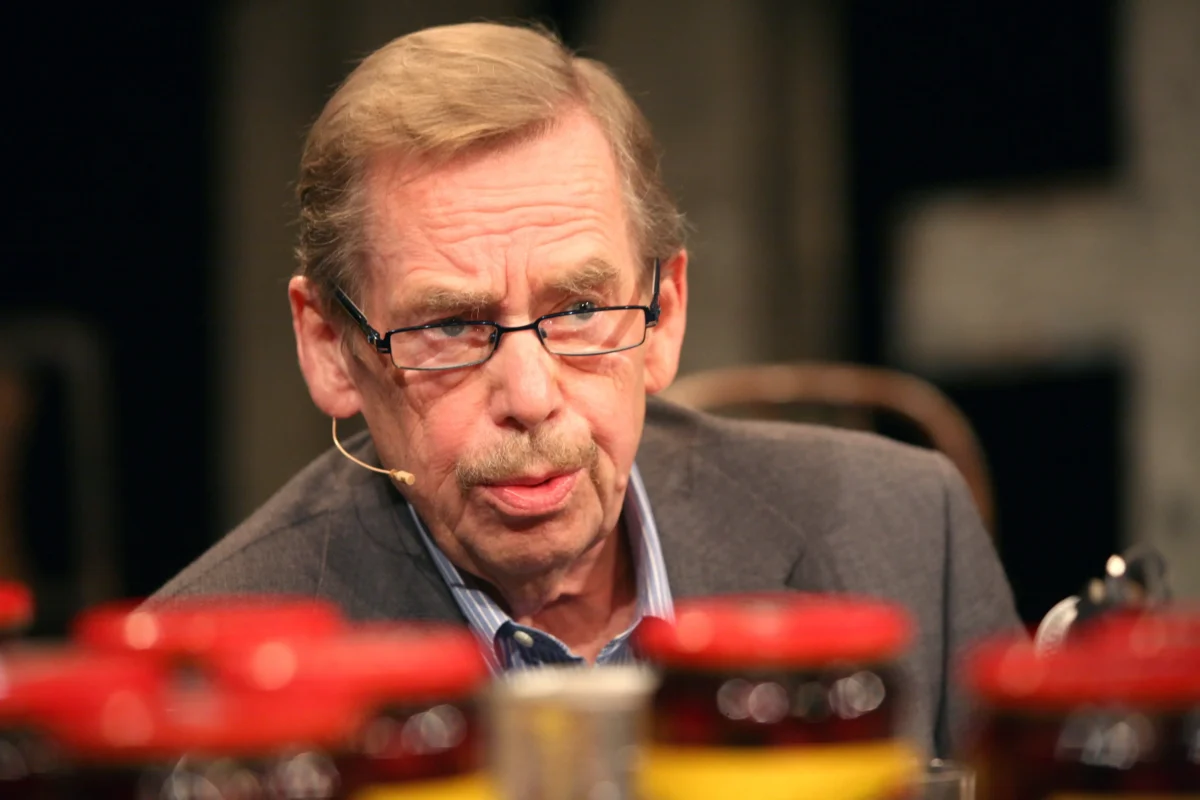 Close-up of an older man wearing glasses and a headset microphone, looking downward, at a panel or discussion.