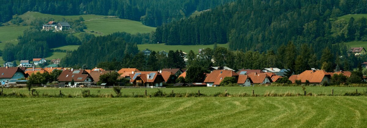 shutterstock_2109351281-změněno-na-3000x1051 | TVGURU.cz Venkovská vesnice s červenými taškovými střechami zasazená podél zeleného pole pod zalesněnými kopci a vzdáleným horským pozadím