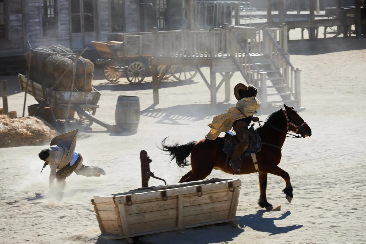 shutterstock_2464495399 1 | TVGURU.cz Two riders chase a horse-drawn parade through a dusty, sunlit western town with wooden storefronts and carts in the background.