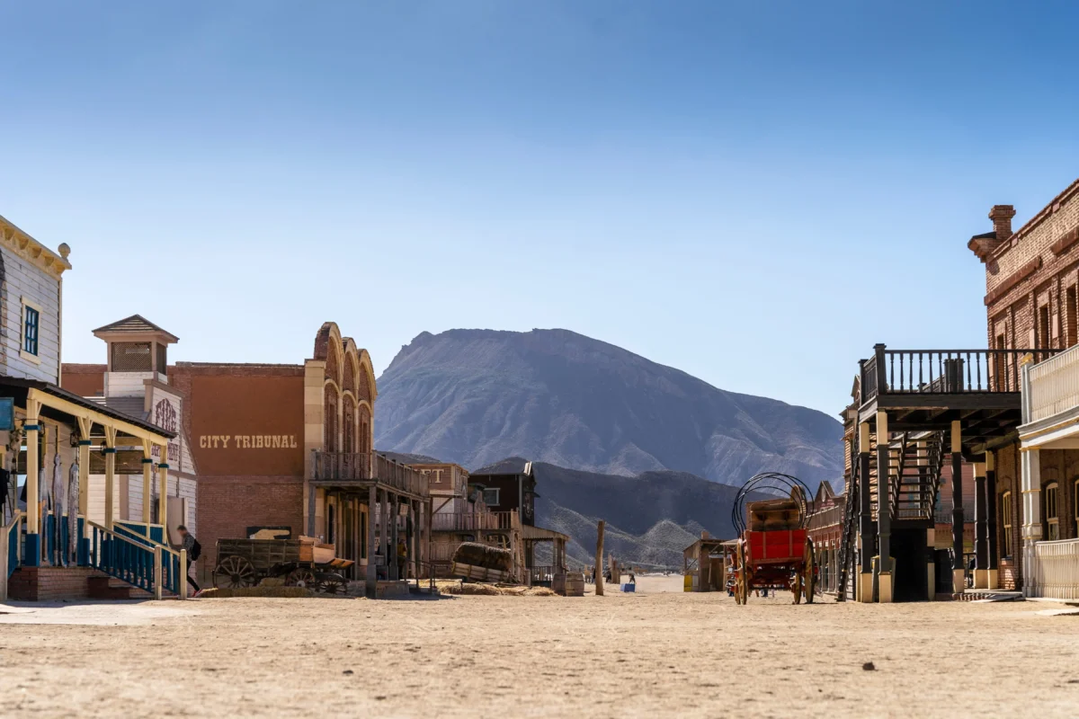 shutterstock_2522063423 1 | TVGURU.cz Desert Western town street with wooden storefronts, mountain in background and clear blue sky; sign reads'CITY TRIBUNAL' on left building
