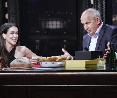 Prostředí televizní talk show s usmívající se hostující ženou a moderátorem u stolu, na stole viditelný chléb, pečivo a nápoje. V popředí s Janem Krausem