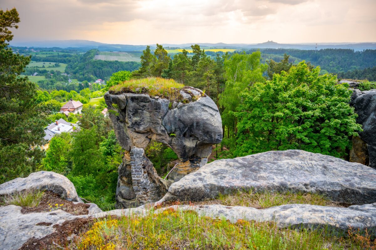 shutterstock_2020195523-změněno-na-3000x2000 | TVGURU.cz Skalnatá vyhlídka s přírodním kamenným obloukem, zelenými stromy a údolím s motýlkem pod ní. Vzdálené kopce pod zataženou oblohou odhalují poklidnou krajinu.