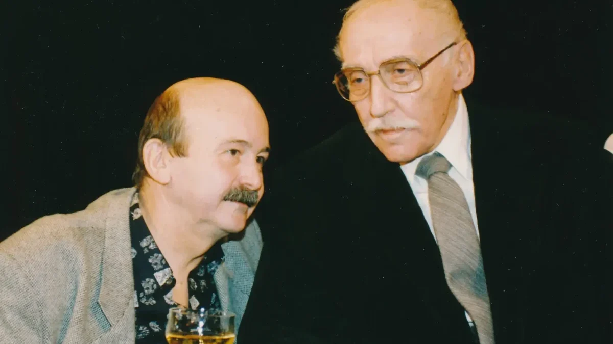 Two older men in a social setting, one in a gray blazer holding a glass of amber drink as they converse.