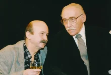 Two older men in a social setting, one in a gray blazer holding a glass of amber drink as they converse.
