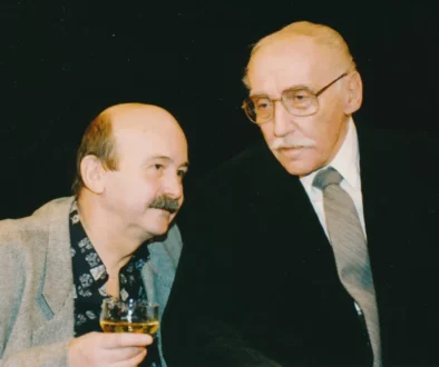 Two older men in a social setting, one in a gray blazer holding a glass of amber drink as they converse.