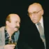 Two older men in a social setting, one in a gray blazer holding a glass of amber drink as they converse.
