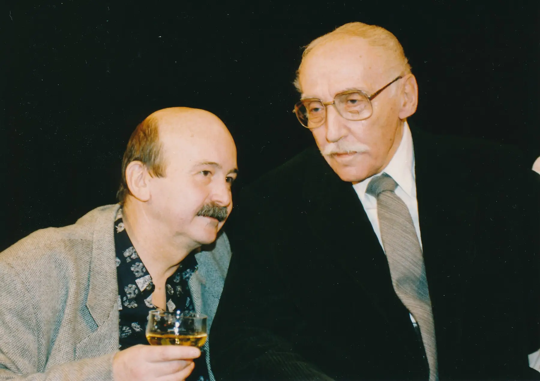 Two older men in a social setting, one in a gray blazer holding a glass of amber drink as they converse.