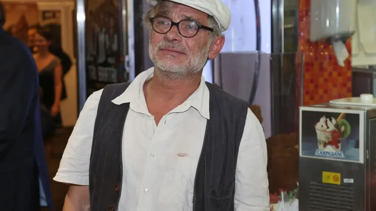 Older man with gray beard and glasses wearing a white cap, white shirt, and dark vest, standing indoors.