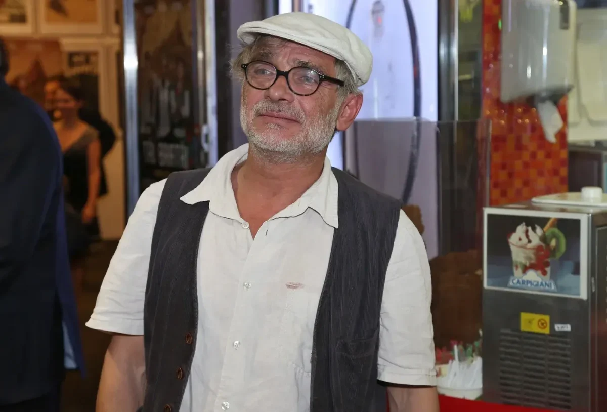 Older man with gray beard and glasses wearing a white cap, white shirt, and dark vest, standing indoors.