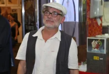 Older man with gray beard and glasses wearing a white cap, white shirt, and dark vest, standing indoors.