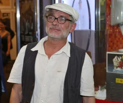 Older man with gray beard and glasses wearing a white cap, white shirt, and dark vest, standing indoors.