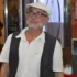 Older man with gray beard and glasses wearing a white cap, white shirt, and dark vest, standing indoors.