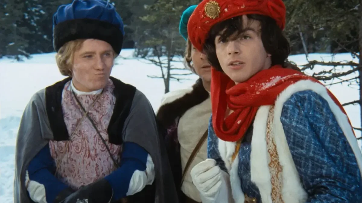 Two costumed men in a snowy outdoor setting, wearing medieval-style robes and hats, looking off to the side.