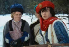 Two costumed men in a snowy outdoor setting, wearing medieval-style robes and hats, looking off to the side.