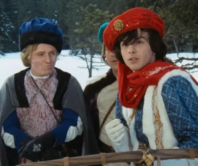 Two costumed men in a snowy outdoor setting, wearing medieval-style robes and hats, looking off to the side.