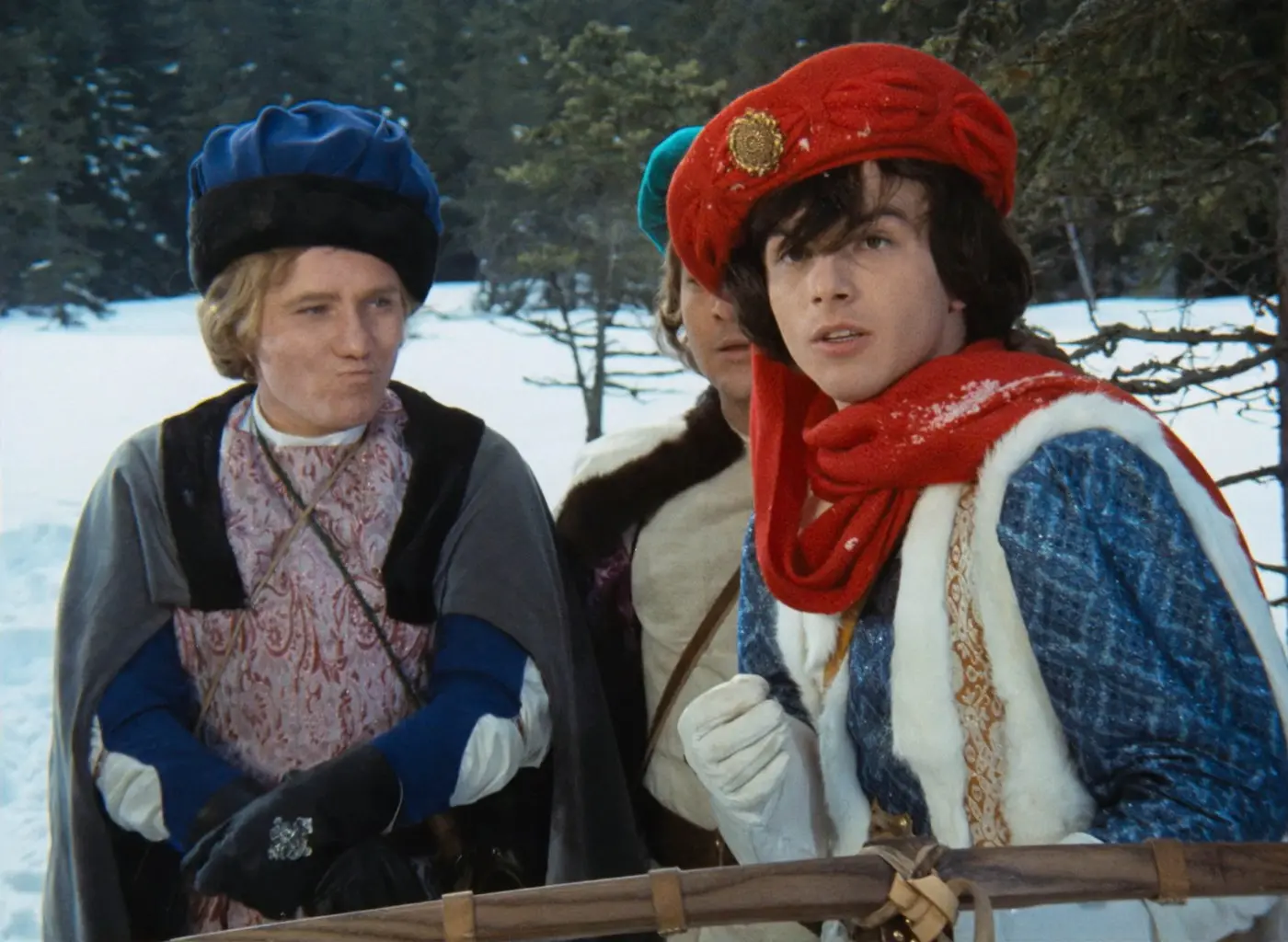 Two costumed men in a snowy outdoor setting, wearing medieval-style robes and hats, looking off to the side.