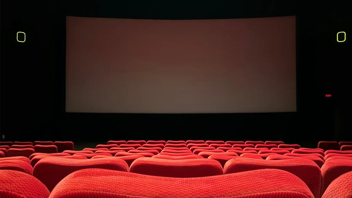 Empty red-velvet theater seats facing a large blank screen on a dark stage.