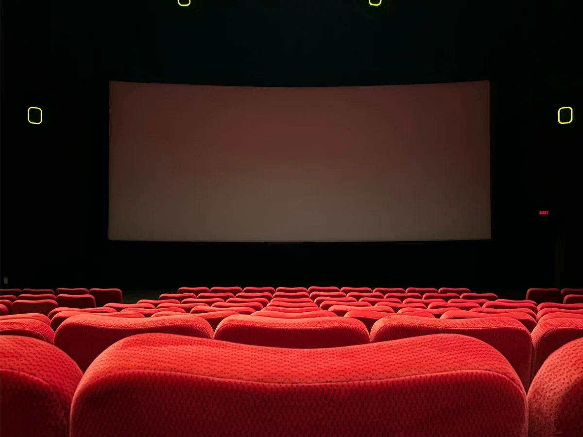 Empty red-velvet theater seats facing a large blank screen on a dark stage.