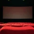 Empty red-velvet theater seats facing a large blank screen on a dark stage.