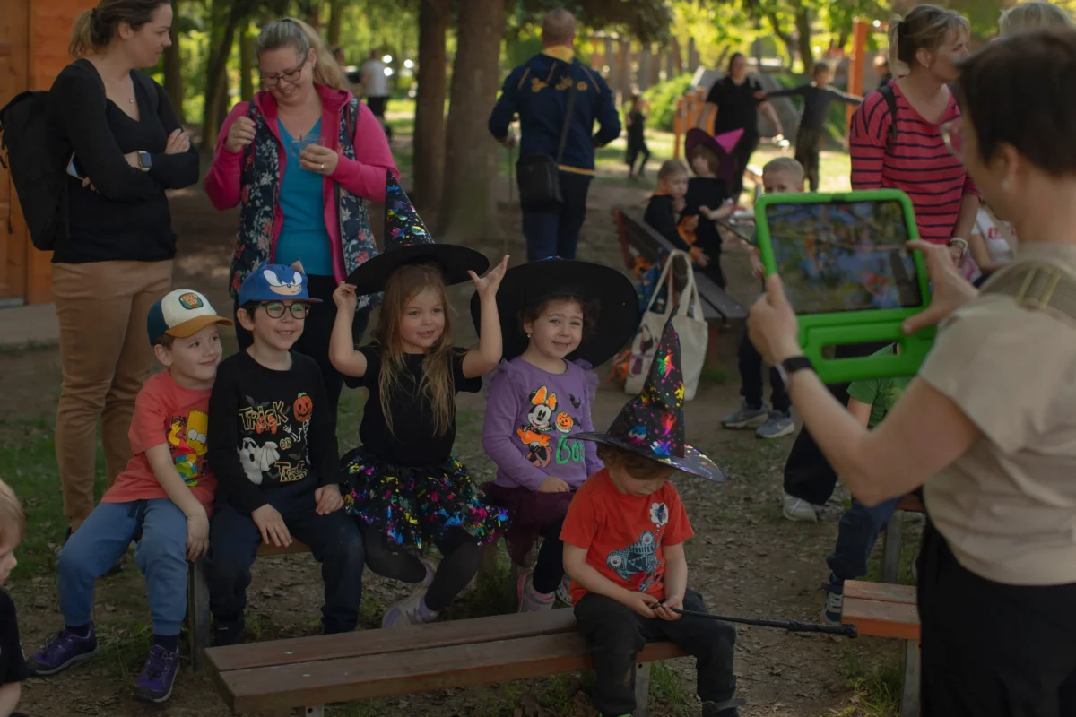 shutterstock_2620422615 1 | TVGURU.cz Group of young children in Halloween costumes posing on a bench in a park while adults watch and a person films in the foreground